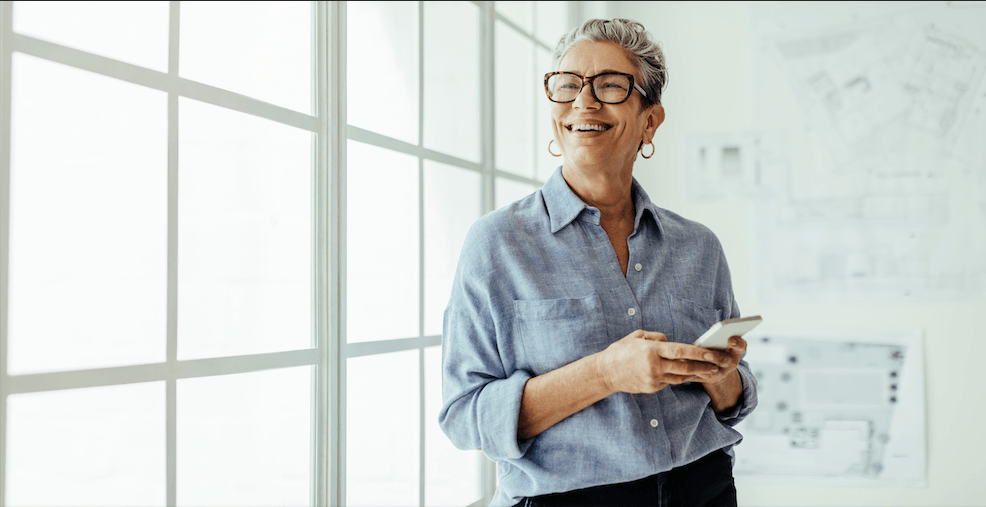 Broker standing near window holding phone and considering benefits strategies for multi-generational workforce